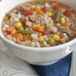 close up image of ground turkey, mixed vegetable and barley soup in a white bowl on a white plate with blue napkin.
