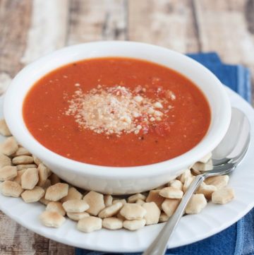 Side view image of Easy Homemade Tomato Basil Parmesan soup in white bowl with oyster crackers and spoon on blue fabric.