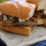 Two halves of a steak and onion sandwich with blue cheese on a hoagie bun on a white plate on a blue napkin.