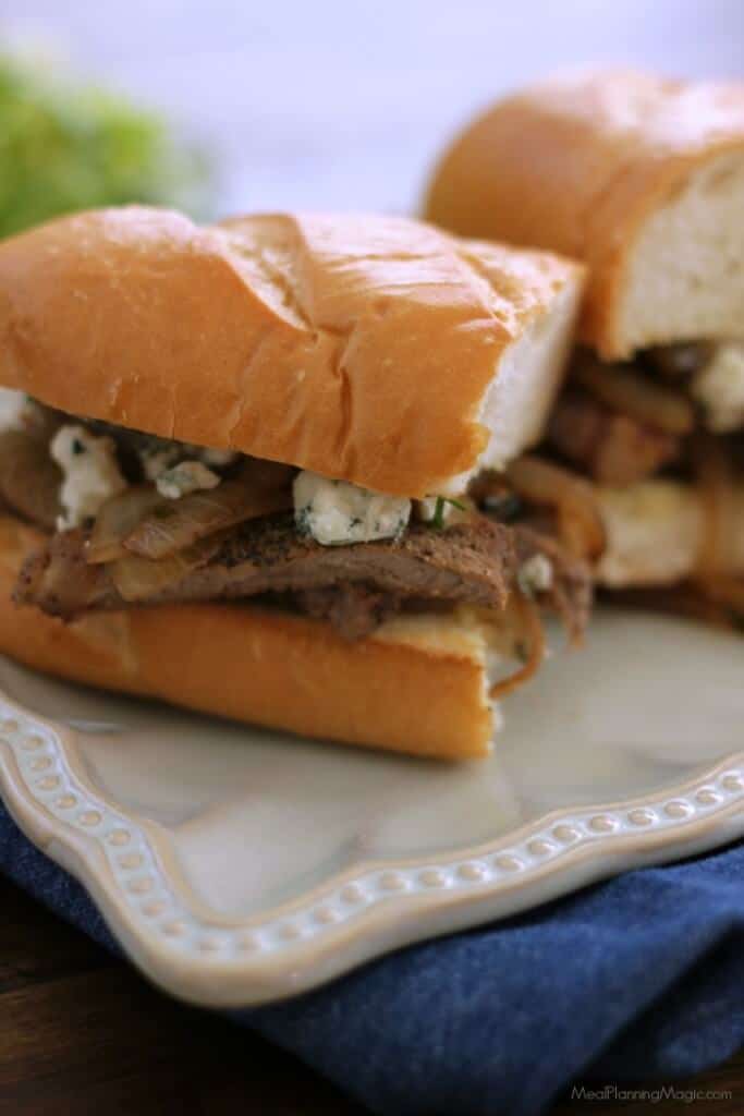 Two halves of a steak and onion sandwich with blue cheese on a hoagie bun on a white plate on a blue napkin. 