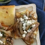 Two halves of an open faced steak and onion sandwich with blue cheese crumbles on top on a square white plate on a blue napkin.