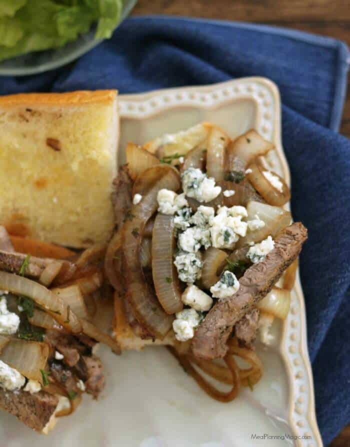 Two halves of an open faced steak and onion sandwich with blue cheese crumbles on top on a square white plate on a blue napkin.