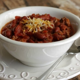 Slowcooker Classic American Chili with beans topped with grated cheddar cheese in a white bowl on a white plate with spoon on side.