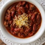 top view image of slowcooker classic American chili with beans topped with shredded cheddar cheese in a white bowl on a white plate