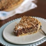 Slice of German Chocolate Sweet Cream Pie on white plate and blue plate with whole pie in background.