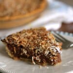 Side view of slice of German Chocolate Sweet Cream pie on white plate on blue plate with whole pie in background.