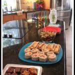 Buffet of different food on platters on a black counter in a kitchen.