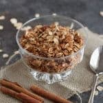 Slow Cooker Honey Cinnamon Granola in a clear bowl on a burlap napkin with cinnamon sticks and spoon on side