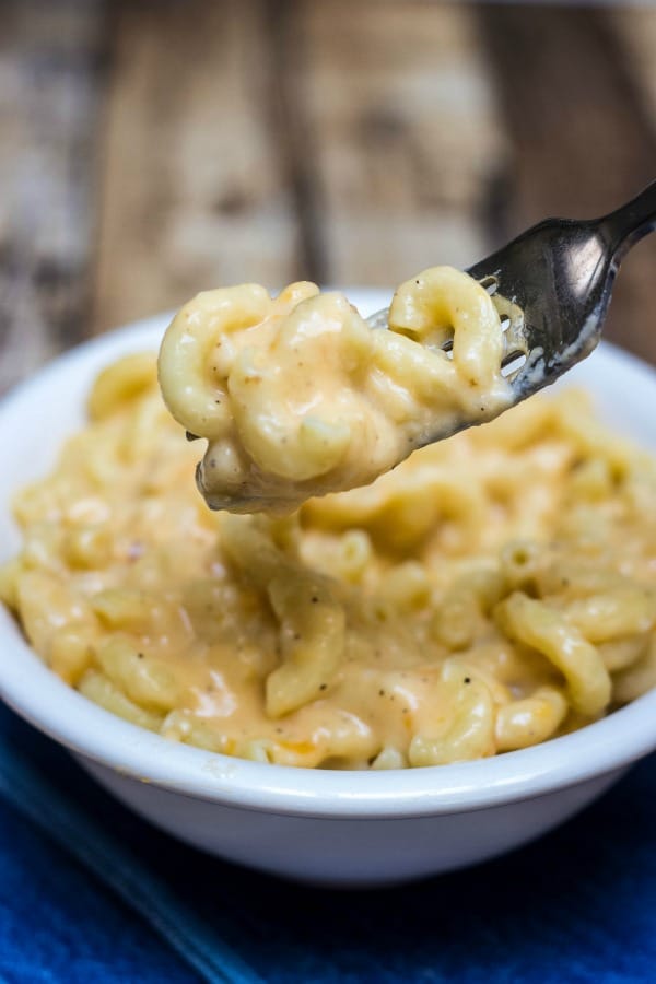 Image of Homemade Macaroni and Cheese in a white bowl on blue napkin with forkful above bowl. 