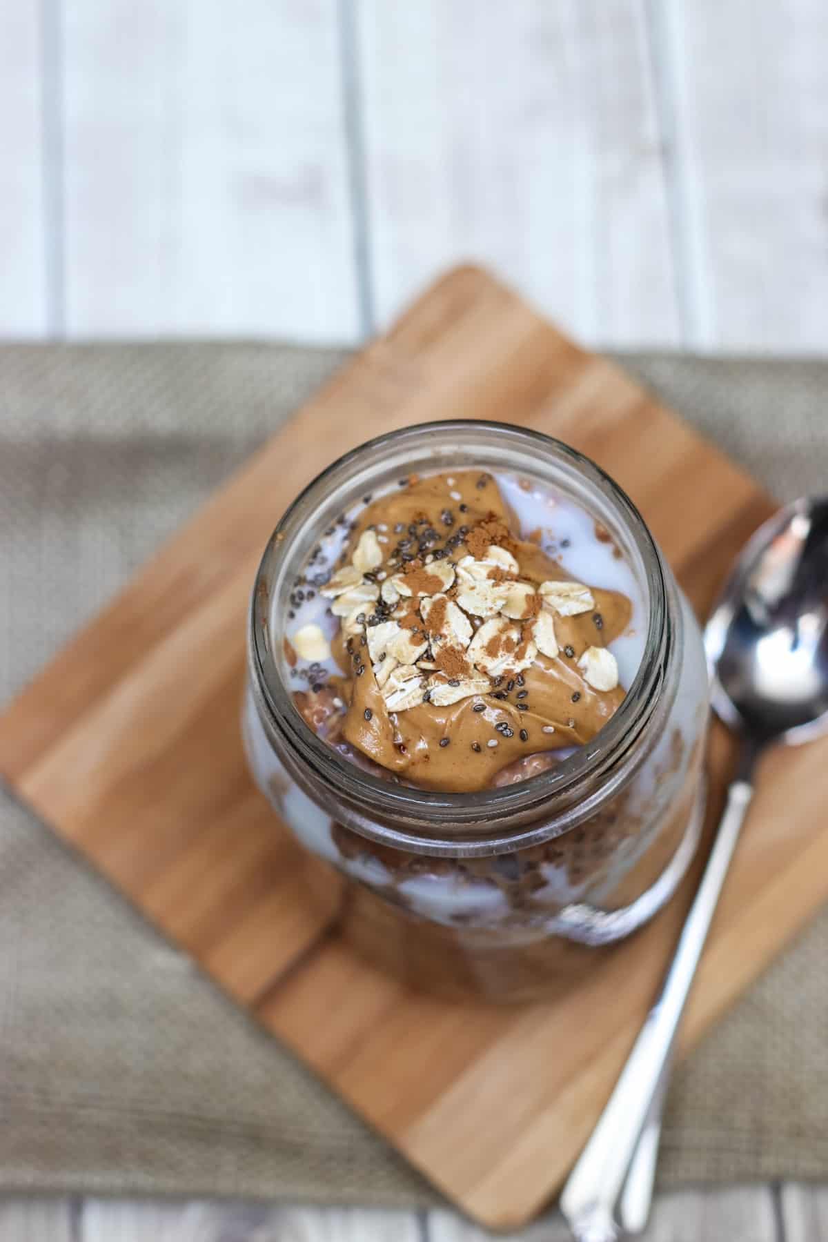 Top view of Cinnamon Chocolate Peanut Butter Overnight Oats in glass jar with peanut butter, oats and chia seeds on top and spoon on side. 
