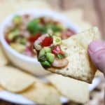 Shrimp Avocado Salsa close up on a tortilla chip with bowlful in background.