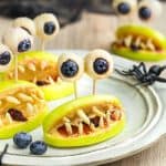 Image of apple slices with slivered almonds to look like teeth and white chocolate dipped blueberries for eyes on a white plate for a Halloween snack.