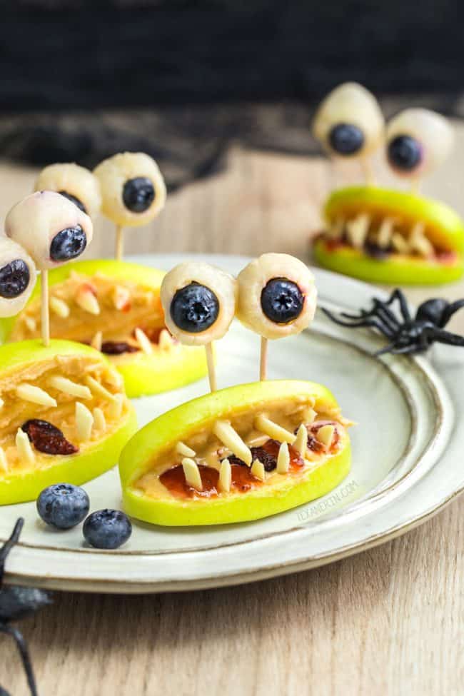 Image of apple slices with slivered almonds to look like teeth and white chocolate dipped blueberries for eyes on a white plate for a Halloween snack.