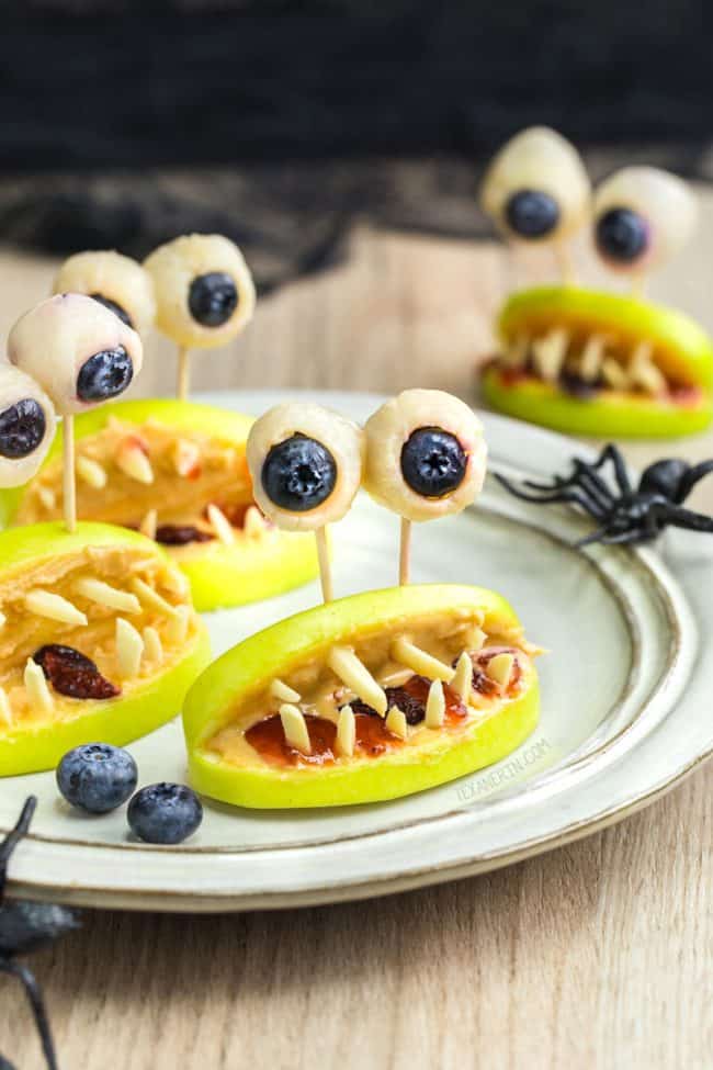 Image of apple slices with slivered almonds to look like teeth and white chocolate dipped blueberries for eyes on a white plate for a Halloween snack. 