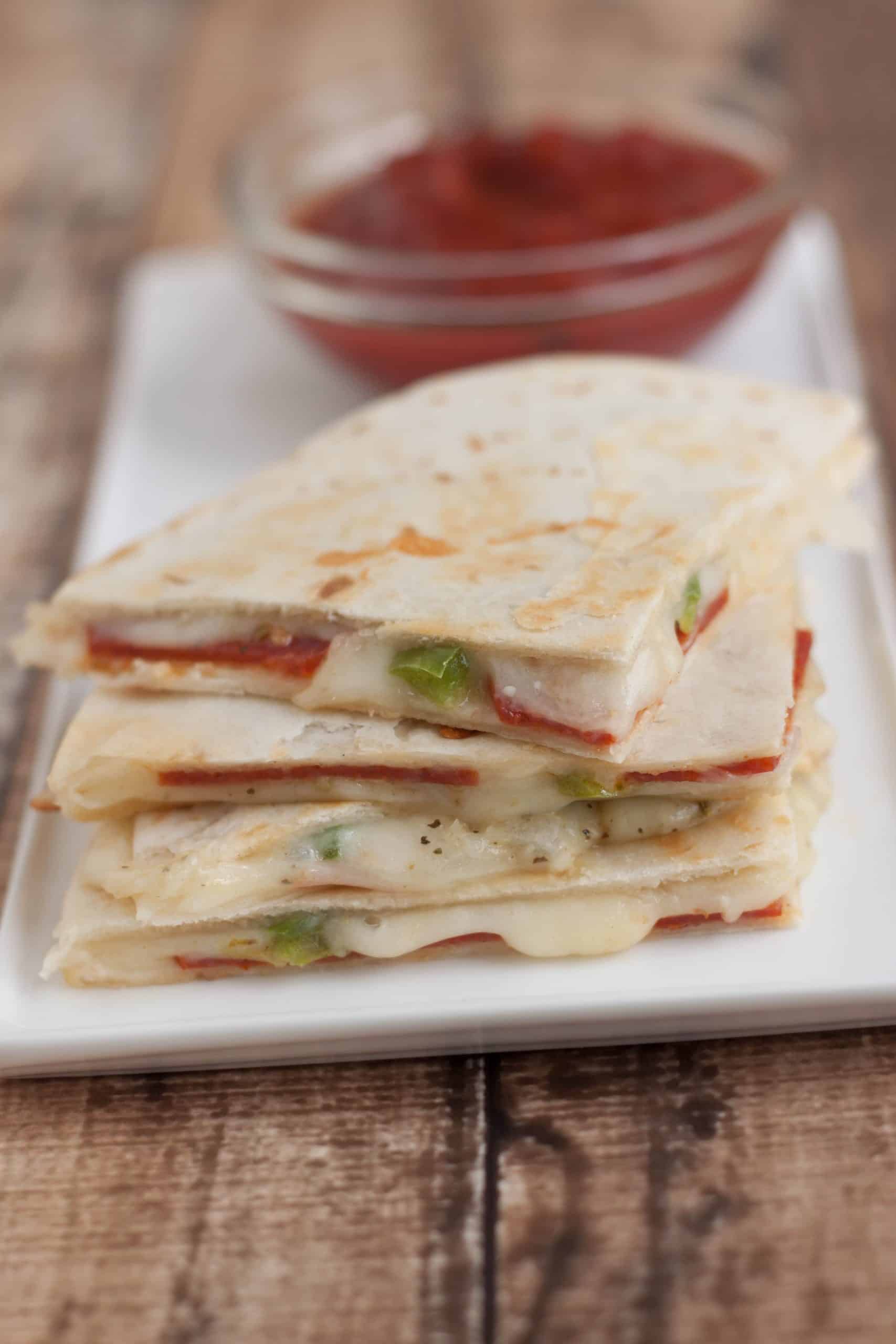 Image of pepperoni pizza quesadilla triangle slices on a white plate with small bowl of pizza sauce for dipping. 