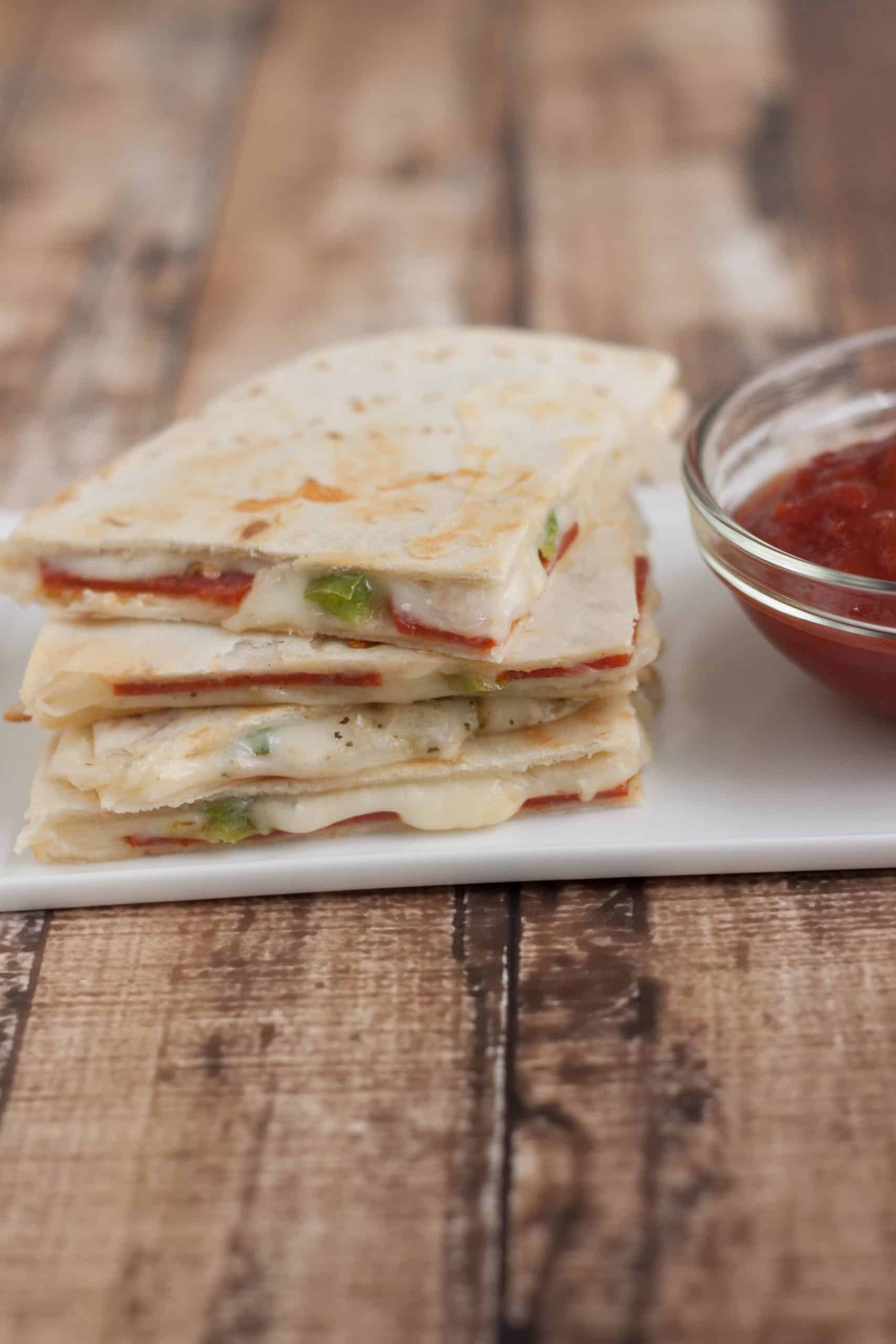 Stack of three pepperoni pizza quesadilla slices on a white plate with pizza dipping sauce on right side.