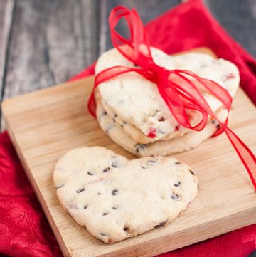 Heart shaped cherry and chocolate chip cookie cutter cookies with three in a stack wrapped in red ribbon on a small wooden cutting board on a red napkin.