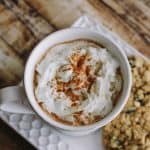 top view image of white mug with whipped cream and cinnamon sprinkled on top with oatmeal cookies on side plate