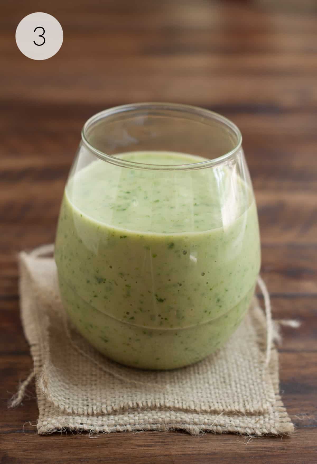 Clear glass filled with banana spinach avocado smoothie on a brown burlap napkin.