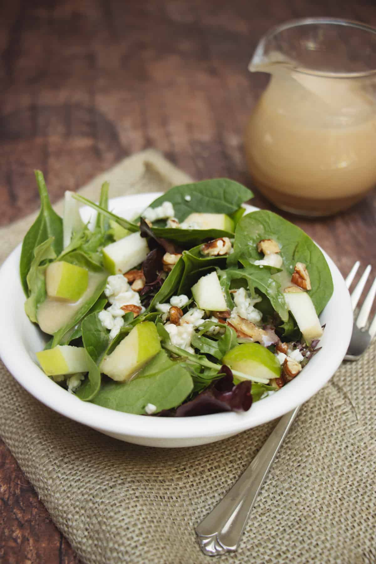 Salad with green apples, blue cheese and pecans in a white bowl on a burlap napkin with dressing in corner.