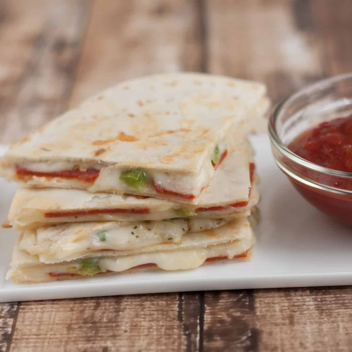 Stack of three Pepperoni Pizza Quesadilla slices with tomato dipping sauce in bowl on a white plate.