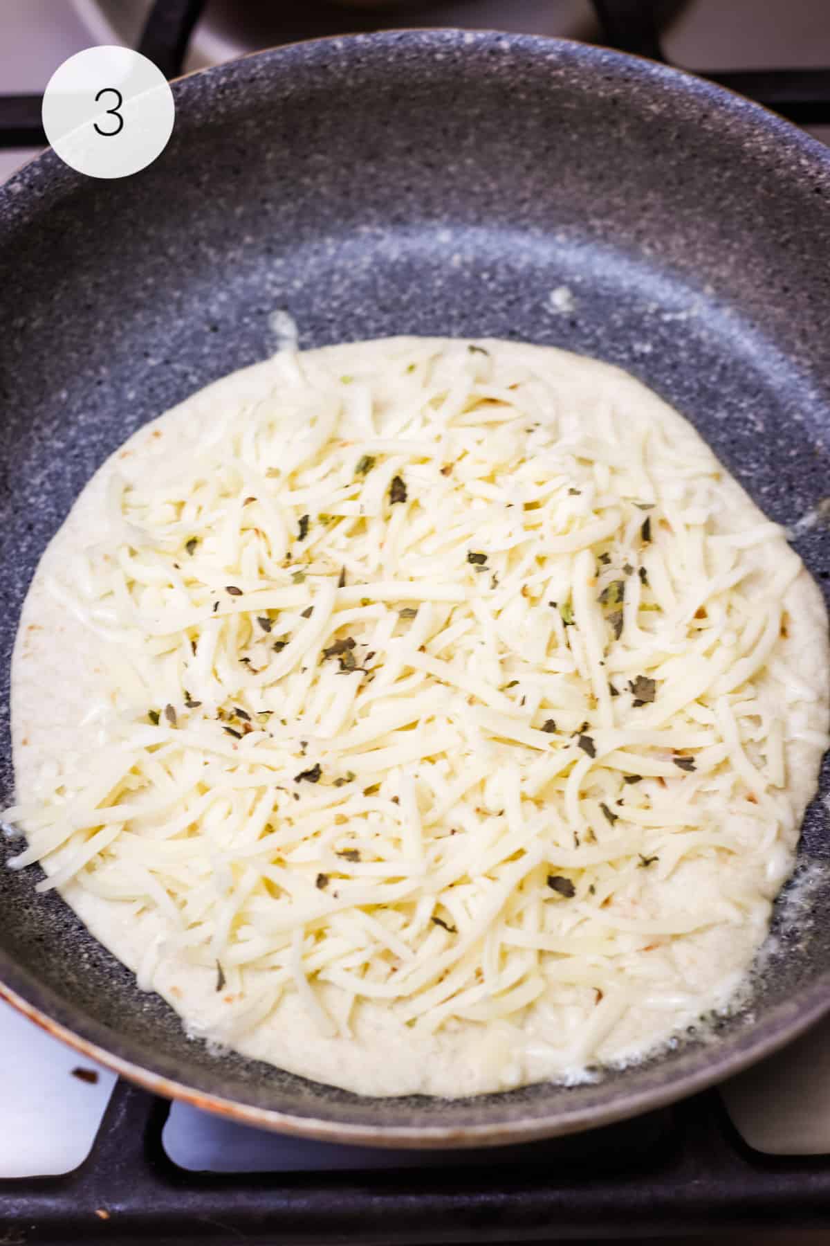 Flour tortilla with shredded mozzarella cheese and herbs cooking in a grey saute pan. 