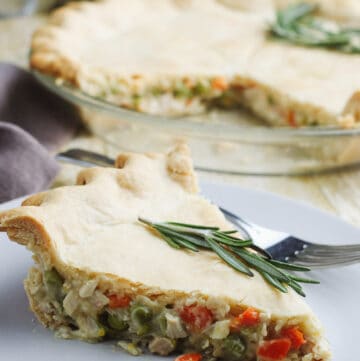 Slice of Homemade Chicken Pot Pie on a plate with rosemary sprig on top and full pie in background.