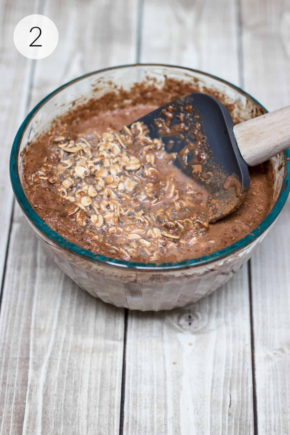 Grey rubber spatula stirring ingredients for chocolate peanut butter overnight oats in a glass bowl.
