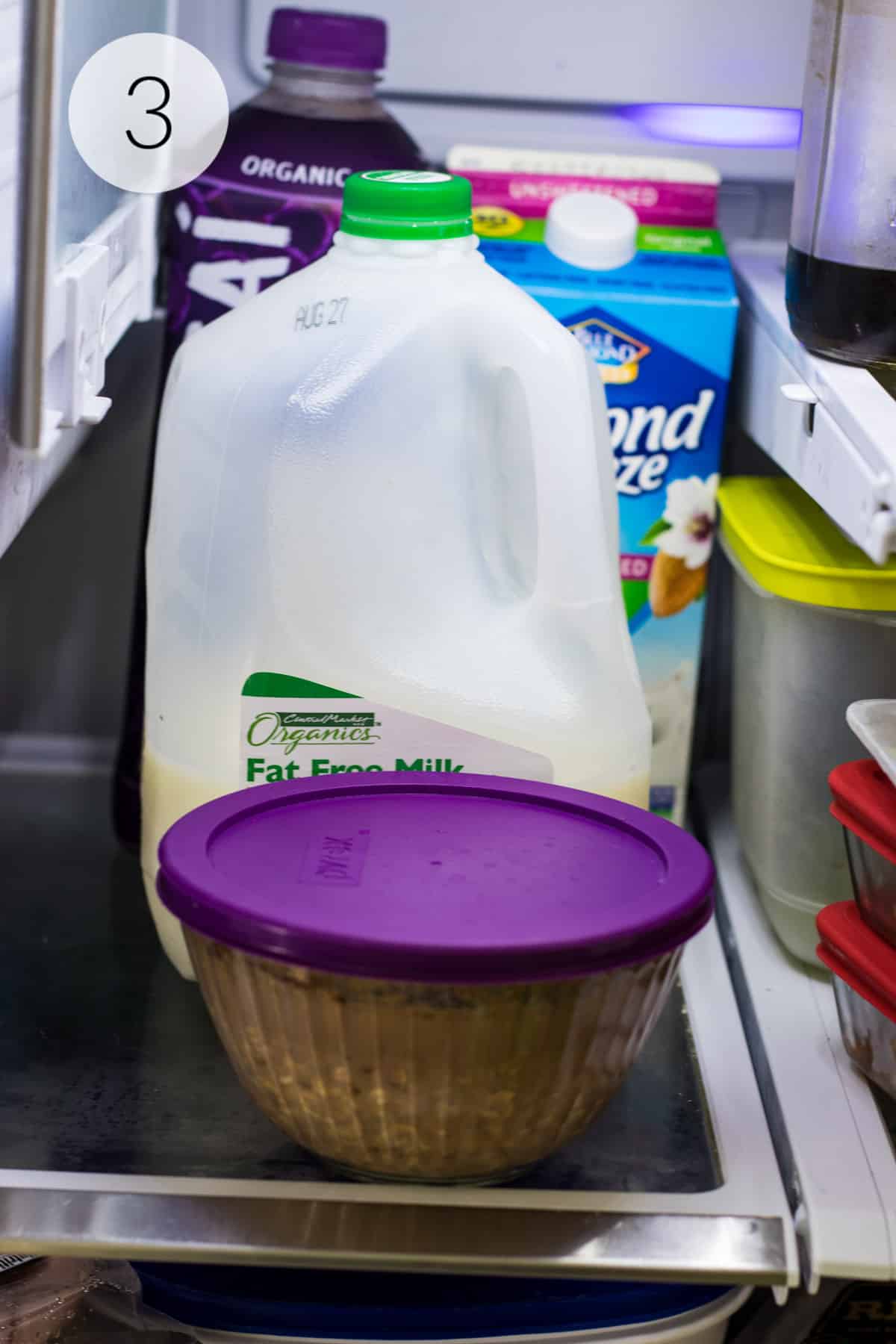 Glass bowl with purple lid containing chocolate and peanut butter overnight oats on shelf in fridge with gallon of milk and other beverages in background.