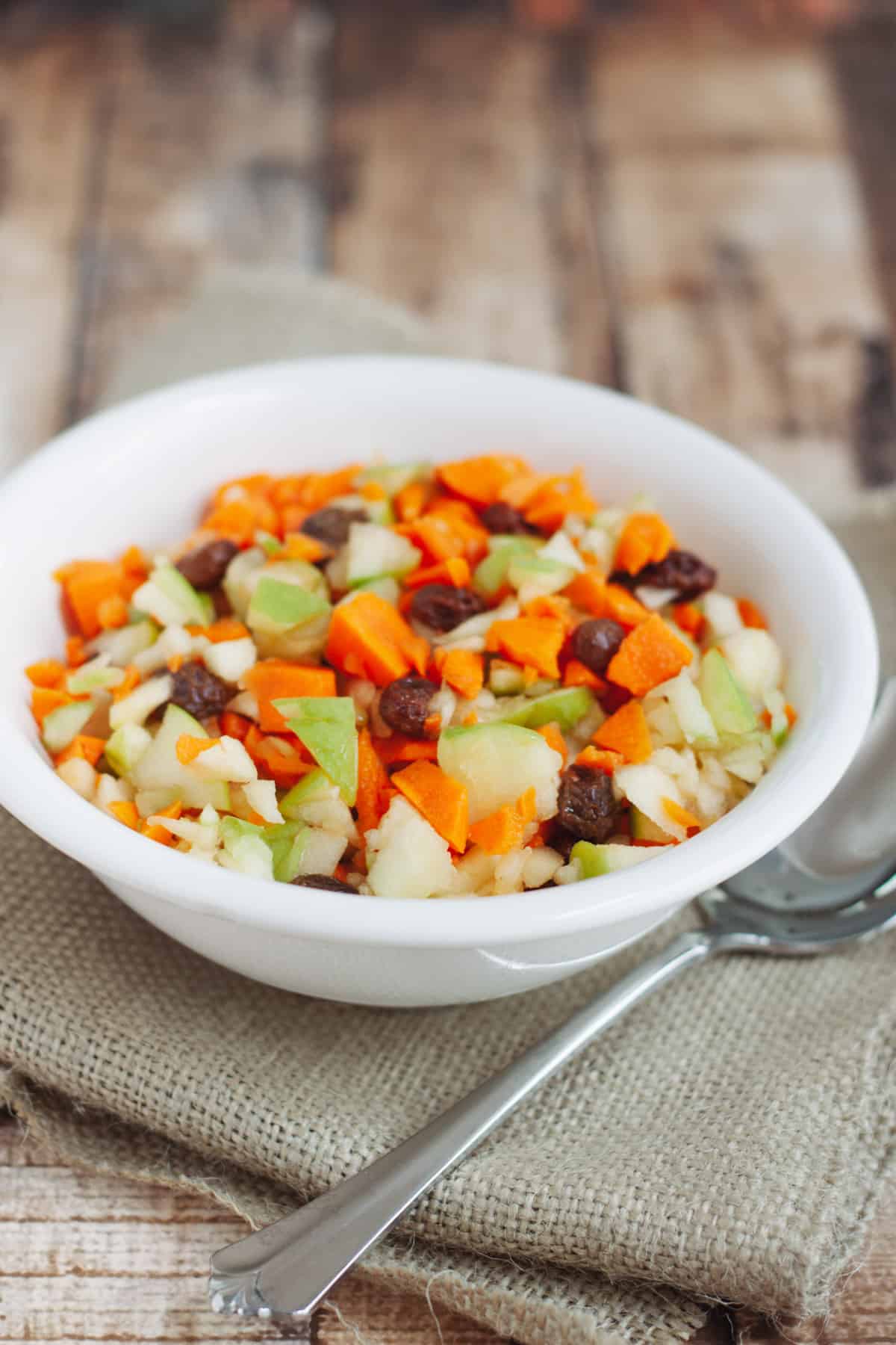 White bowl with finely chopped apples, carrots, and raisins on a burlap napkin with silver spoon. 