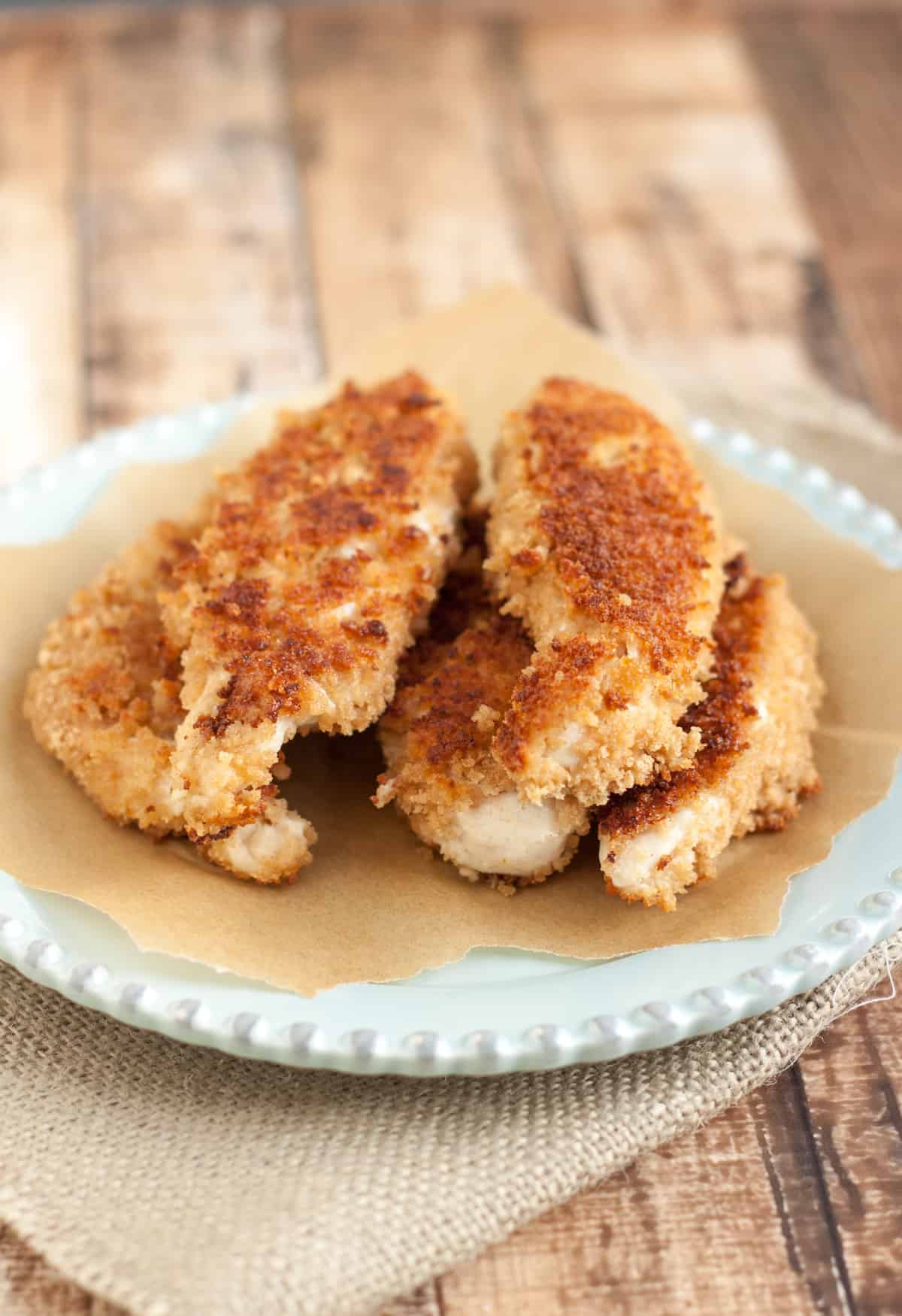 Stack of coconut crusted chicken tenders on a blue plate with parchment paper and burlap napkin.