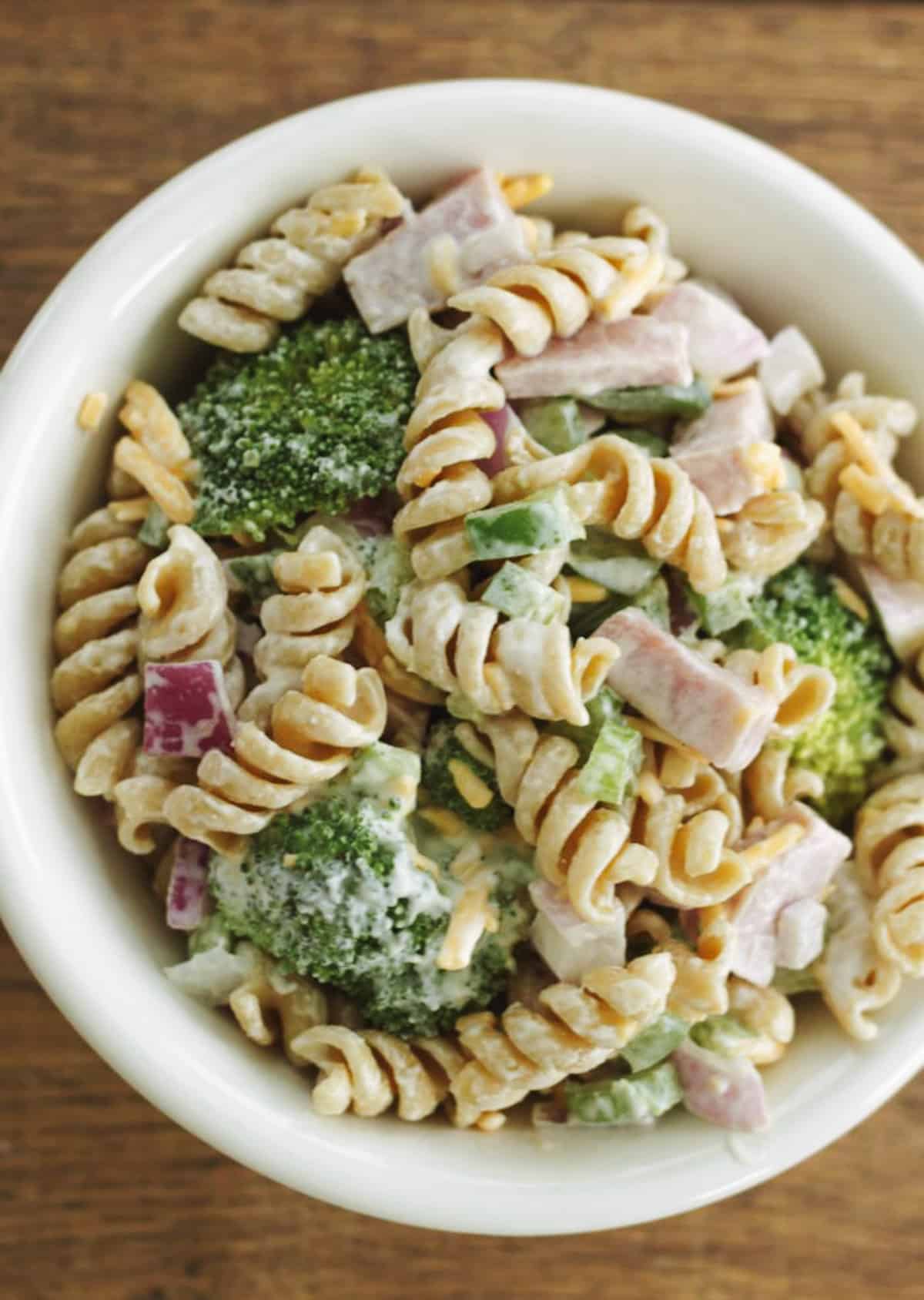 White bowl with pasta salad including broccoli, ham, fusilli pasta, purple onion, cheddar cheese. 