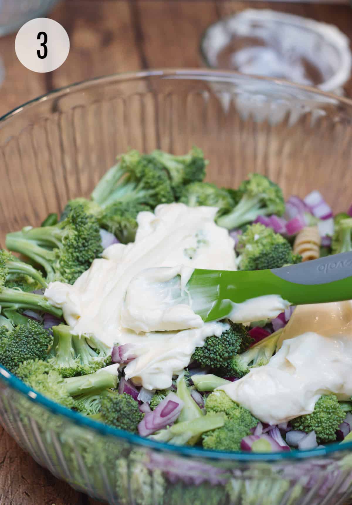 Glass bowl with layered broccoli ham and cheese pasta salad and mayonnaise being spread by green rubber spatula. 