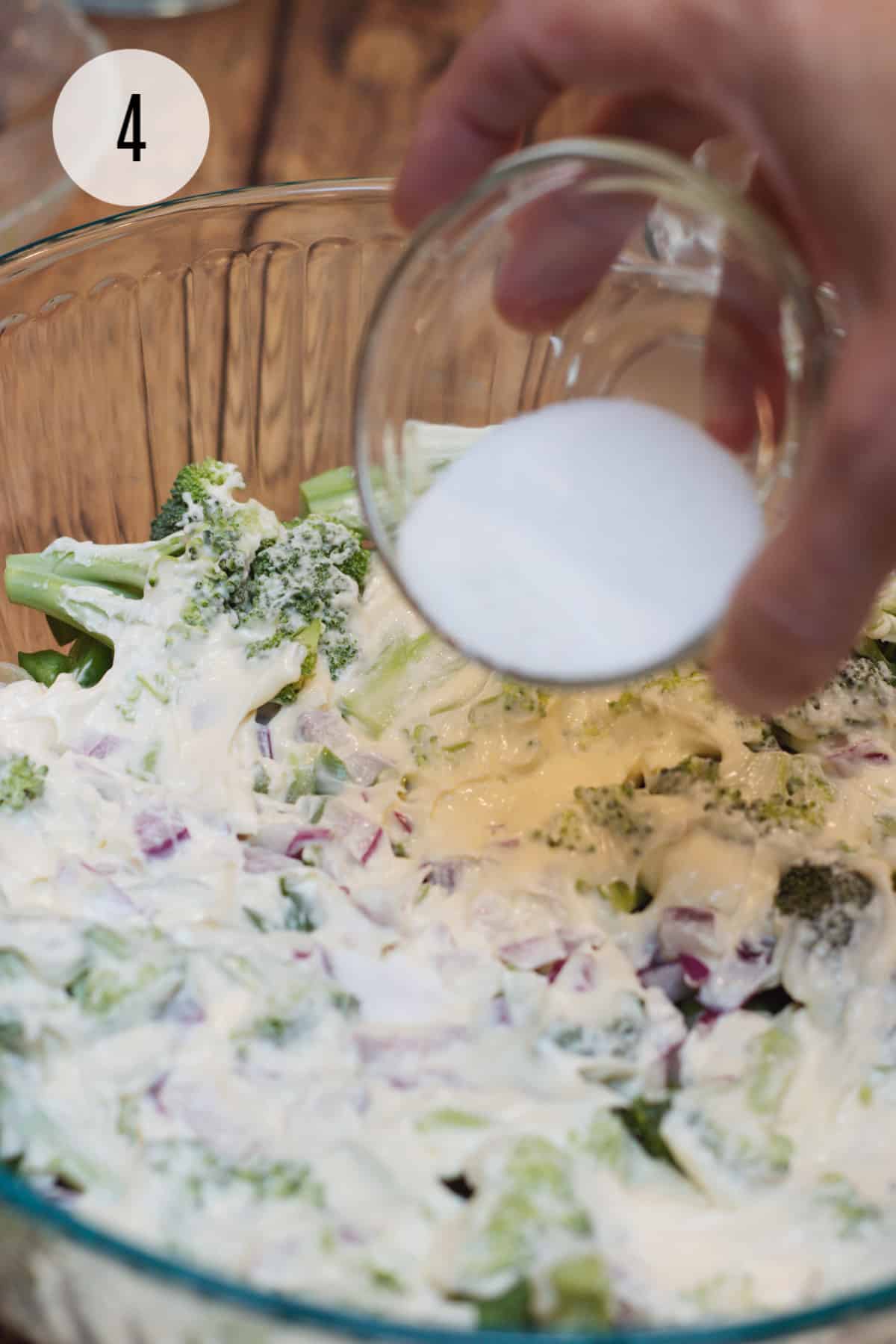 Small bowl of sugar being sprinkled on mayonnaise for broccoli ham and cheese pasta salad in clear bowl. 