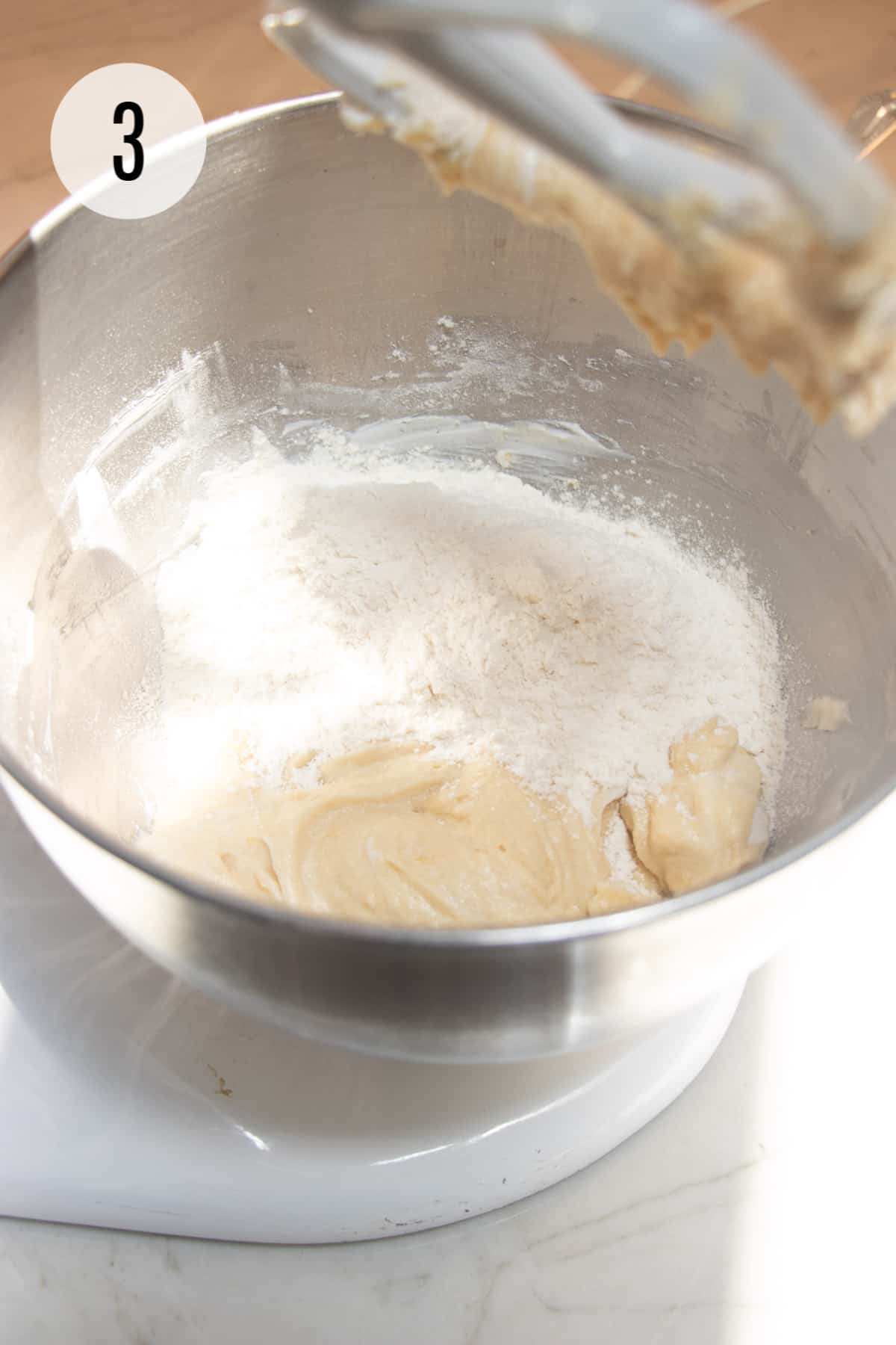 Lemon cookie batter with flour added to a silver bowl mixed by a white mixer paddle.