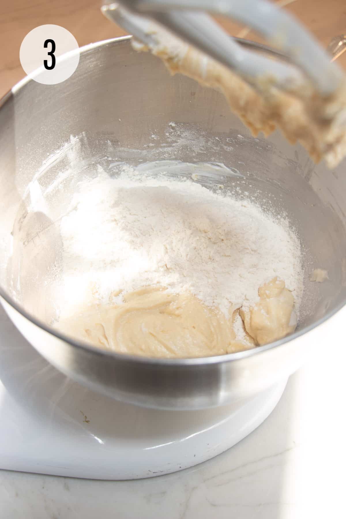 Lemon cookie batter with flour added to a silver bowl mixed by a white mixer paddle. 