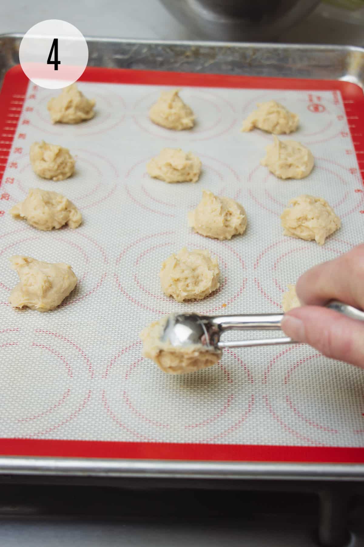 Scoop dropping cookie dough on to a red and white silicone baking mat with cookie dough balls on tray.