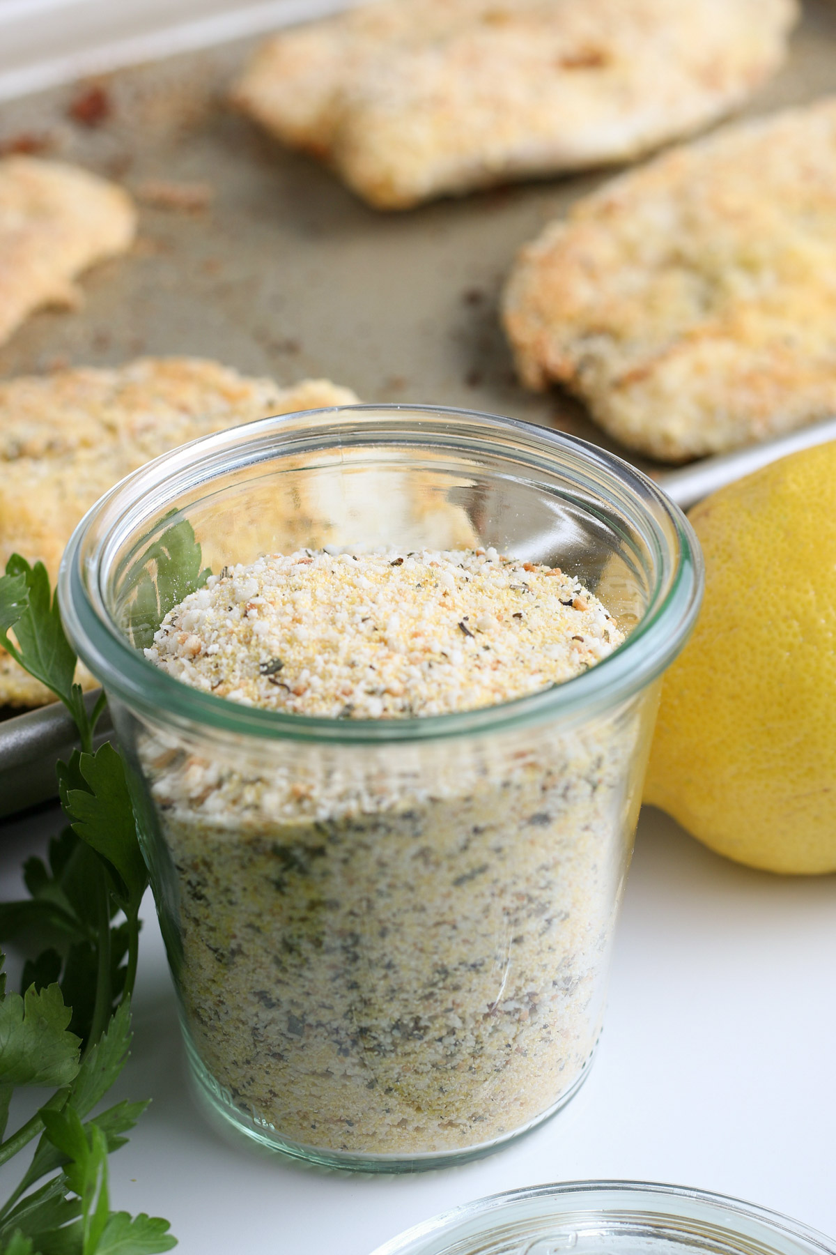 Glass jar of breading mixture for crunchy oven fried breaded tilapia with lemon, parsley and breaded fish in background.