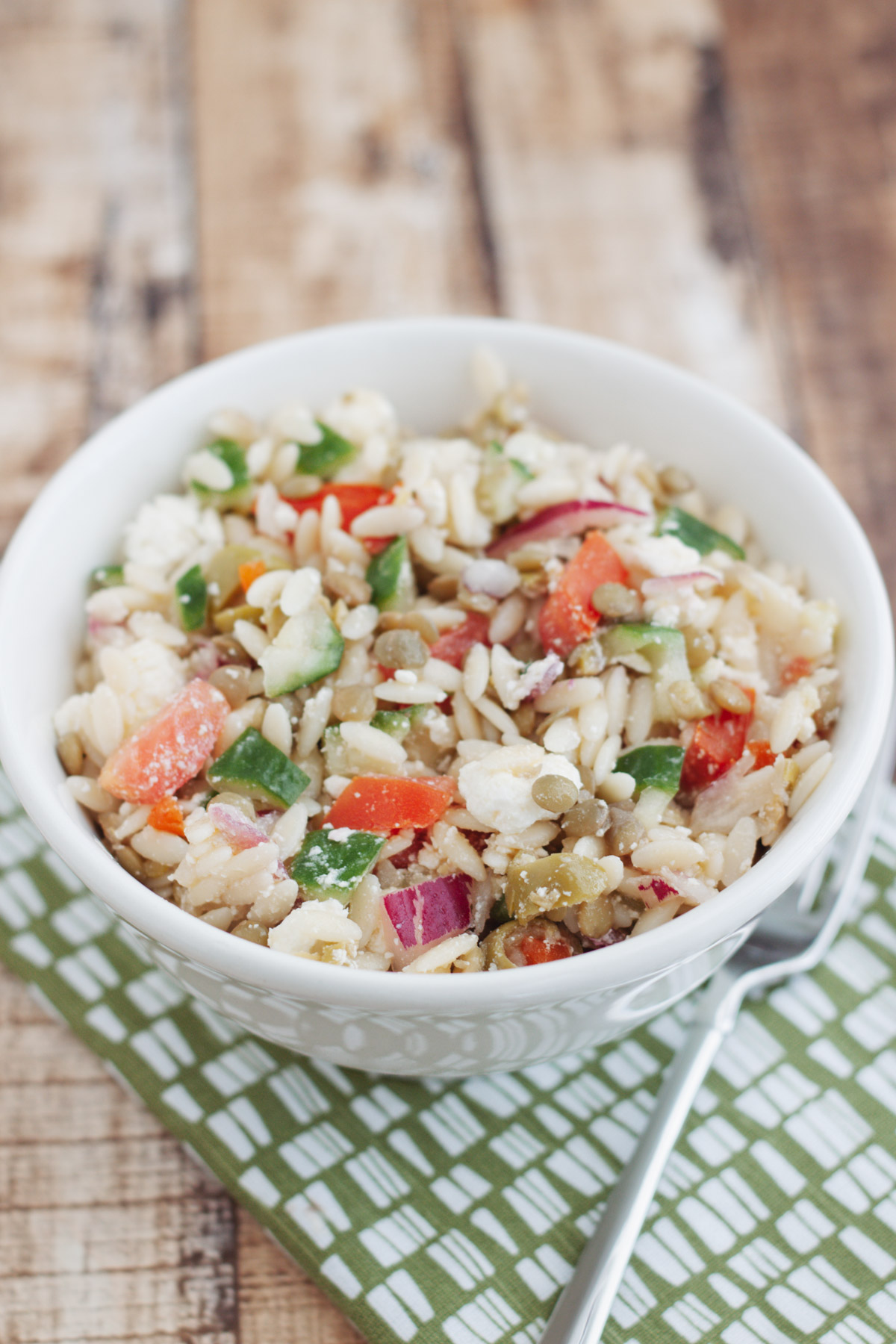 White bowl of Greek Orzo Salad with Lentils and fresh tomatoe and cucumbers in a white bowl on a green and white napkin.