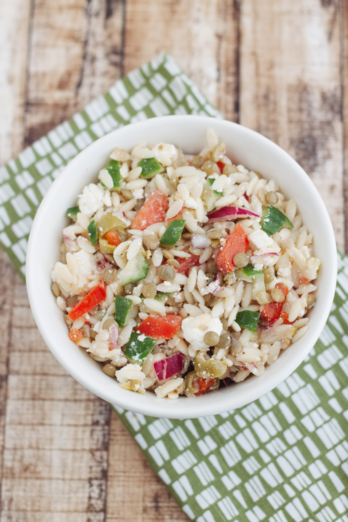 Top view image of Greek Orzo Salad with Lentils in a white bowl on a green and white napkin. 