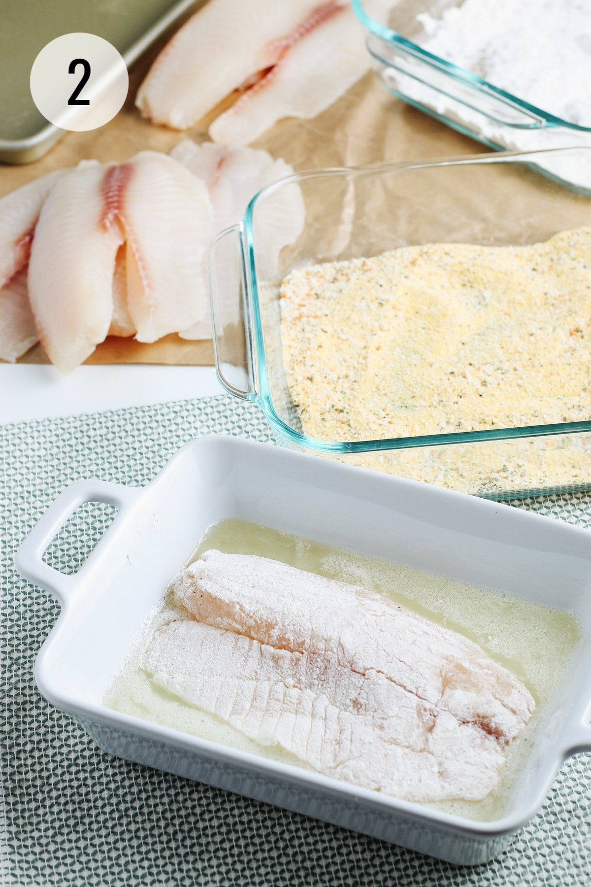 Floured fish filet being dipped in dish with egg whites and extra dish with cornmeal mixture and extra filets placed above. 