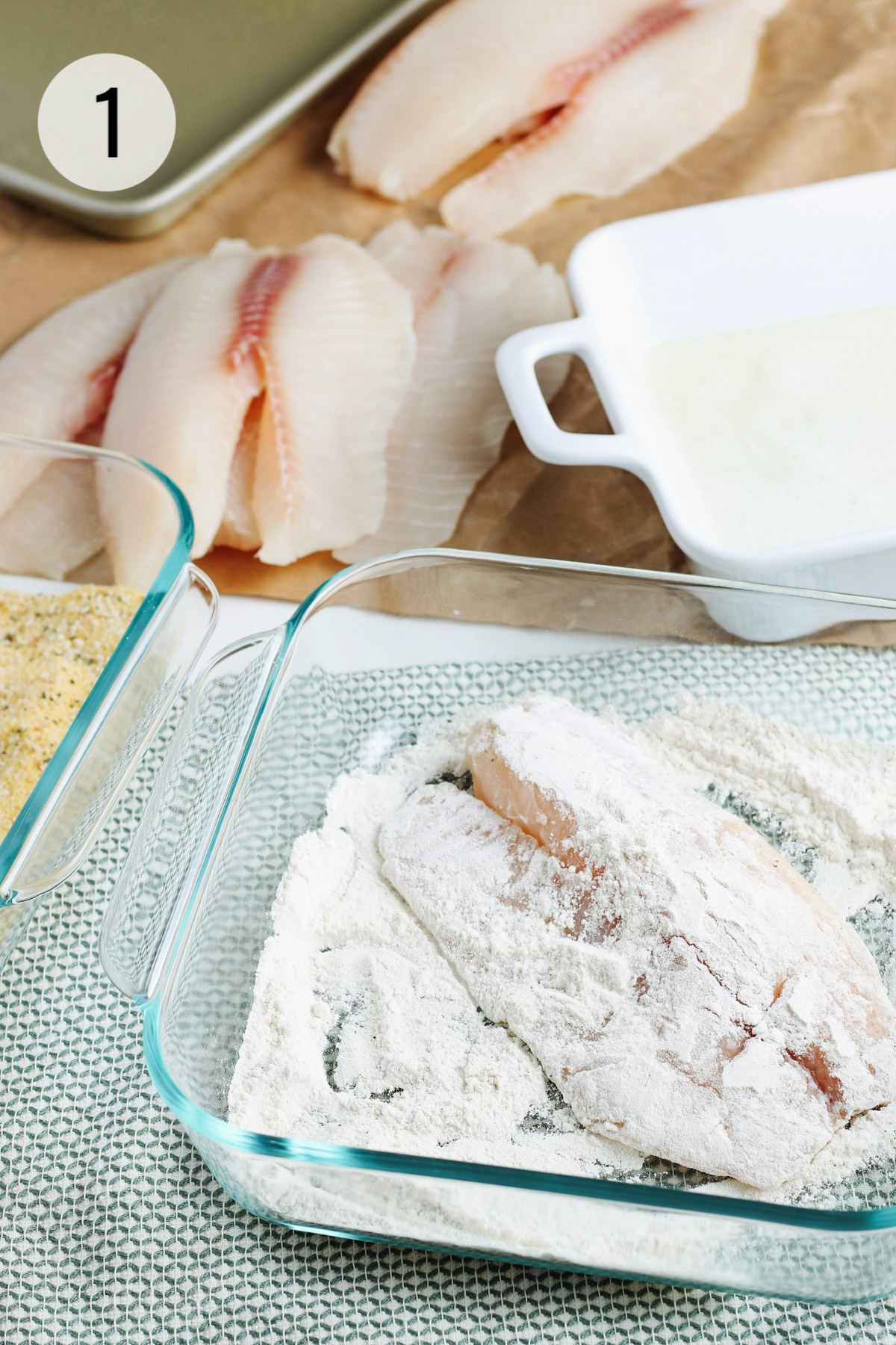 Glass dish with tilapia fish dipped in flour with extra fish filets plus white dish with egg mixture for next step of breading.