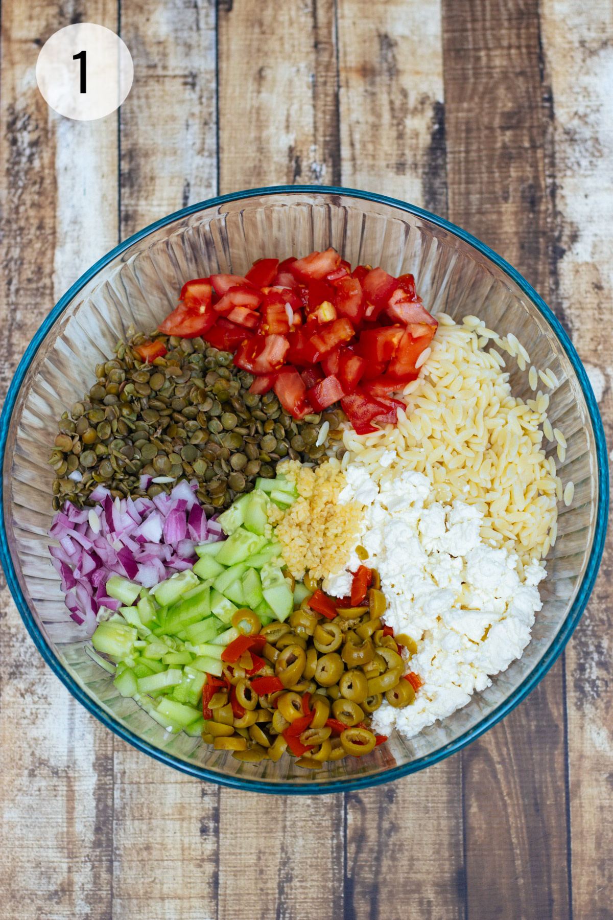 Clear bowl with ingredients for Greek Orzo Salad with Lentils, olives, tomatoes, cucumbers, feta and purple onion. 