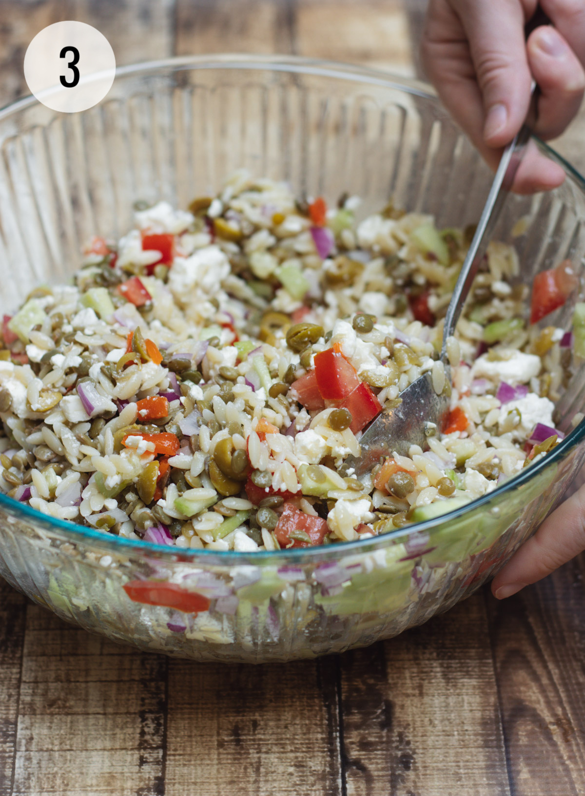 mixing up ingredients for a Greek orzo salad with lentils and vegetables in a clear bowl. 