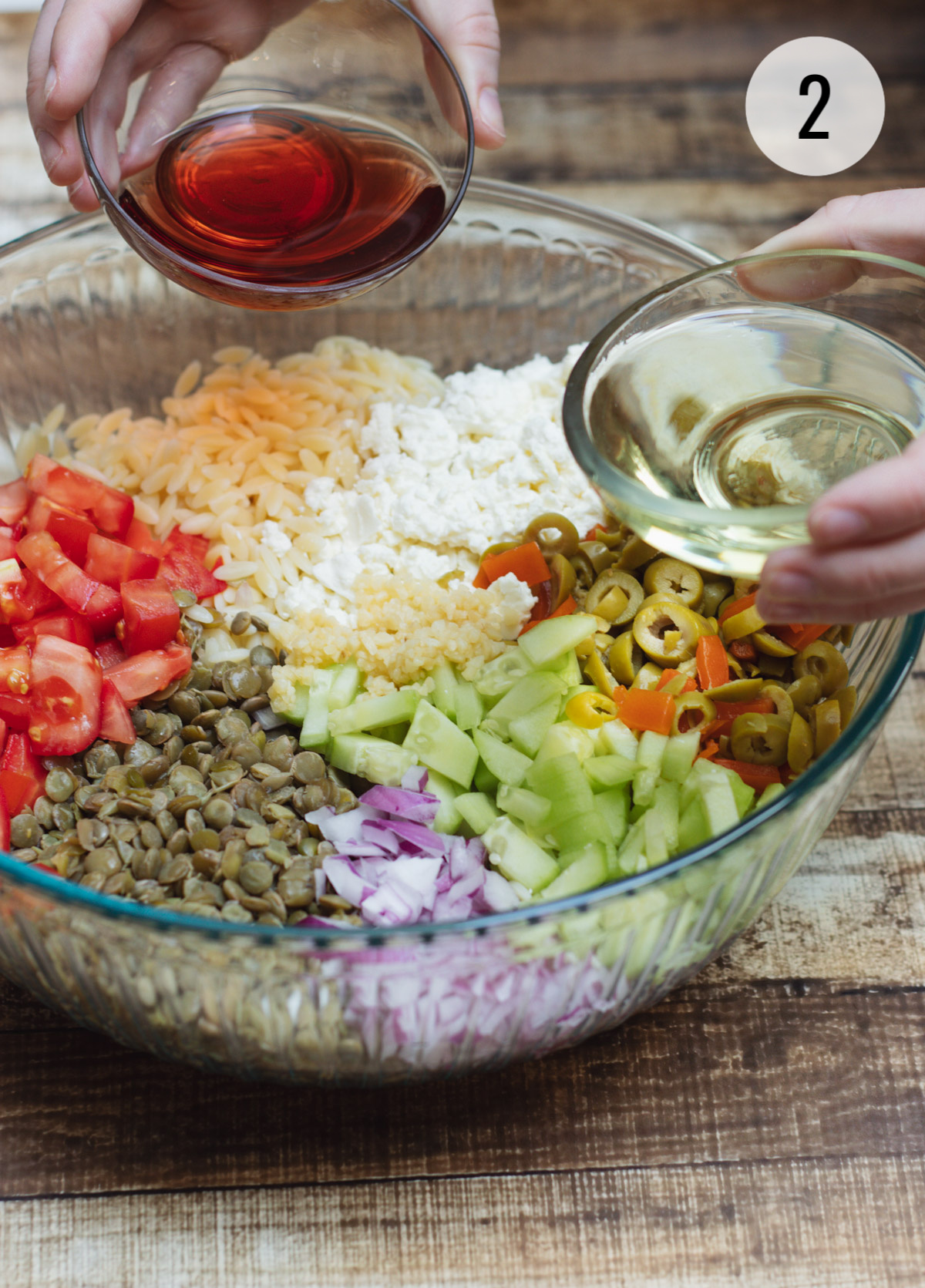 Ingredients for a Greek Orzo Salad with Lentils in a clear bowl with olive oil and red wine vinegar about to be poured in. 