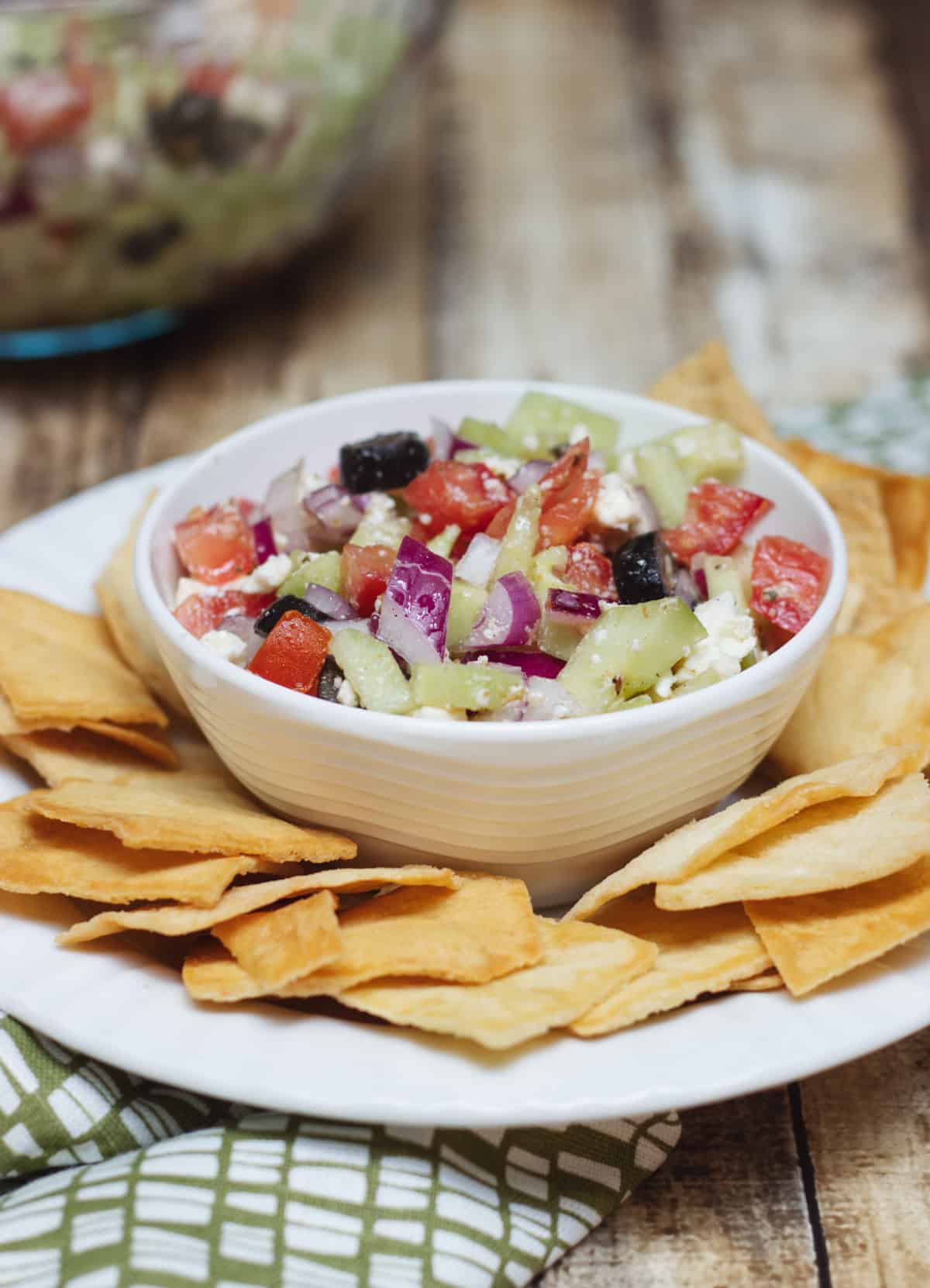 Greek salsa with tomatoes, purple onion, black olives, cucumber and feta cheese in a white bowl surrounded by pita chips.