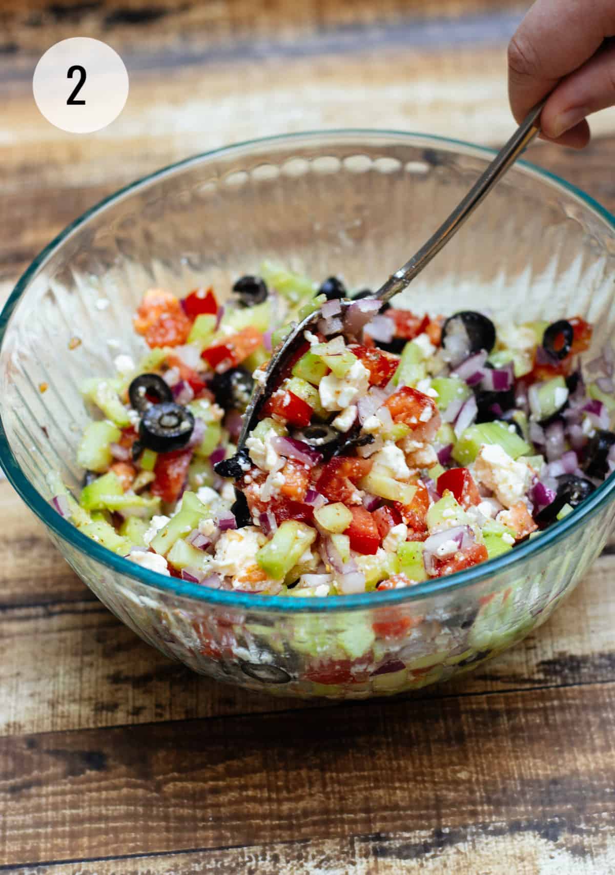 Glass bowl with black olives, tomatoes, cucumbers, feta cheese, and purple onions being stirred by silver spoon.