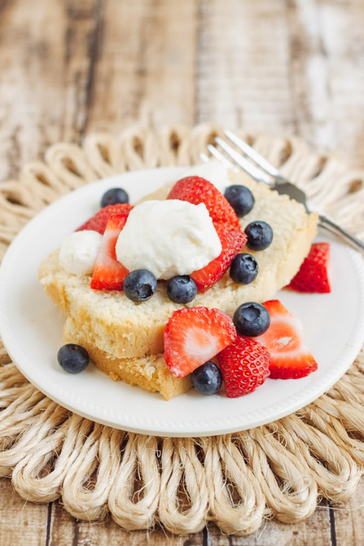 Slices of pound cake on a white plate and straw place mat with strawberries, blueberries and whipped cream on top. 