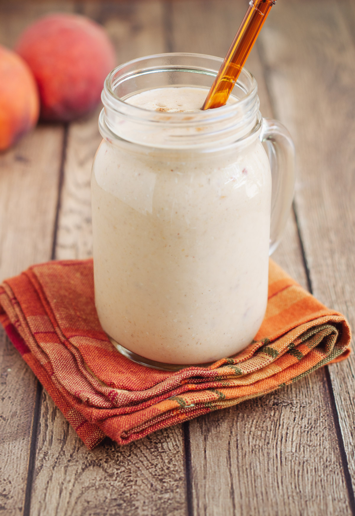 Glass mug with peach smoothie on an orange plaid napkin with orange glass straw in glass and peaches in upper left background.