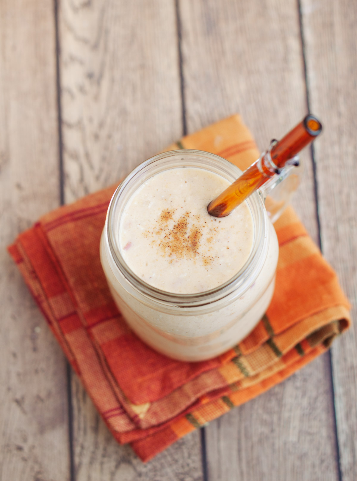 Top view of a peach smoothie with yogurt sprinkled with a bit of cinnamon on a orange plaid napkin and orange glass straw in glass.